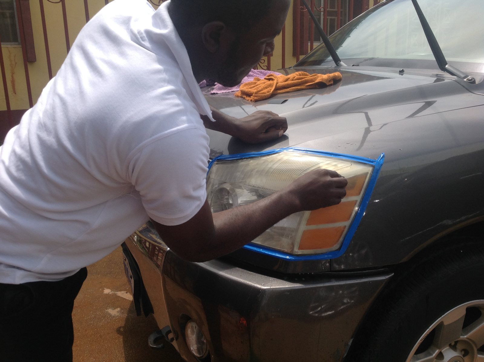 Technician hand-sanding a taped-off yellowed headlight on a vehicle.