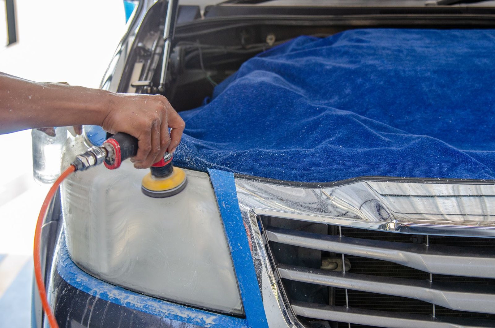 Technician machine-polishing an oxidized headlight protected by masking and a fender cover.